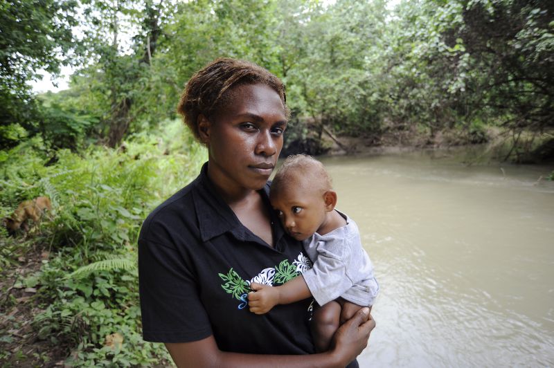 Photo: Lara McKinley/OxfamAUS Portrait of Solomon Islander woman Jemimah John, 21 and her daughter by their river and next to the local church. Jemimah was one of four Solomon Islander women who attended a Women and Mining Conference.
