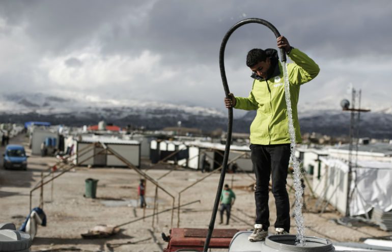 A Syrian refugee and Oxfam volunteer fills a water tank with a hose in a refugee camp, Lebanon