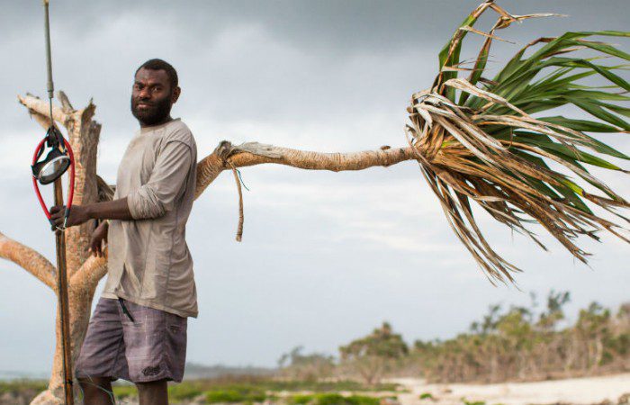 Efate, Vanuatu: Daniel stand on a beach holding a fishing spear and leaning on a withered palm tree.