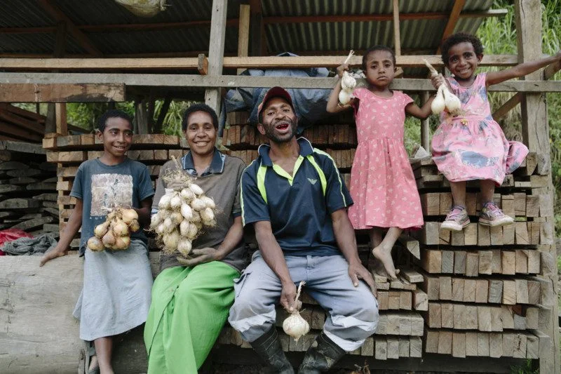 Stephen Bare, with wife Maria Steven, and children *Wendy, *Magen (far right) and *Esther, sit on building materials they will construct their new house from, bought with onion profits. A family sit on building supplies holding onions