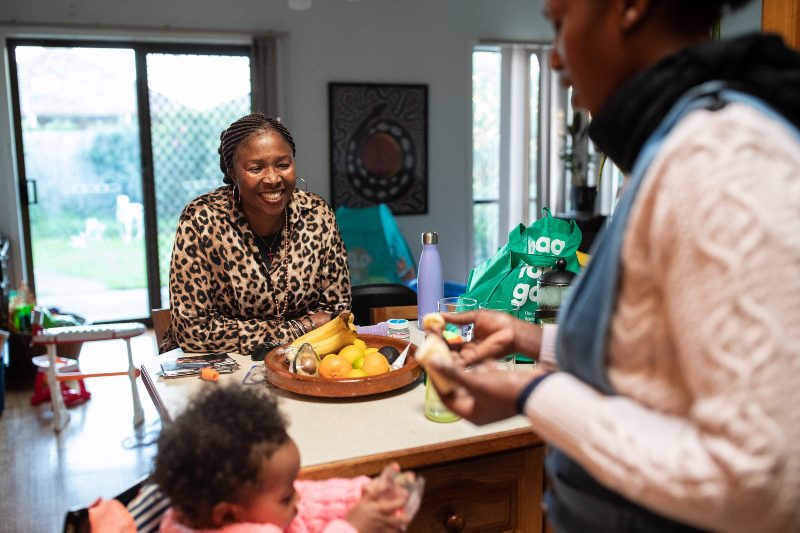 Melbourne, Australia: Lucy (L) and her daugher Susan (R) prepare lunch for *Sophia at Susan’s home in Melbourne. Photo: Kim Landy/OxfamAUS A family sits around a table in a kitchen.