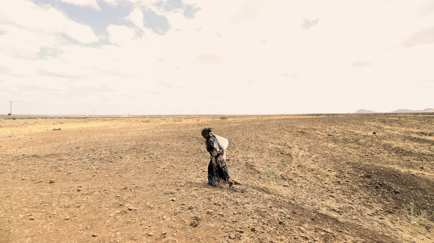 A woman walks across a a desert plain carrying a sack of building stones on her back, determined to rebuild her home. Too many people like her around the world experience poverty fuelled by inequality.