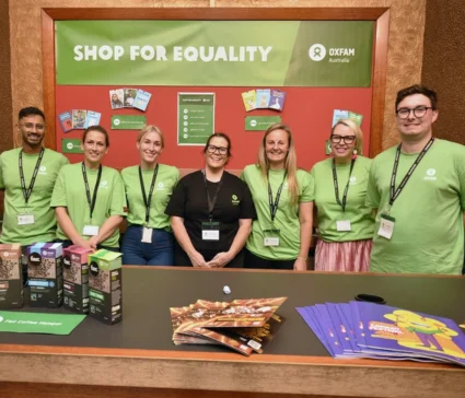 A group of Oxfam volunteers stand in front of an Oxfam banner reading 'Shop for Equality'