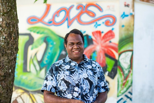 Yannick stands in front of a wall of street art with the word love. Yannick is the program manager of Vanuatu Pride (VPride).
