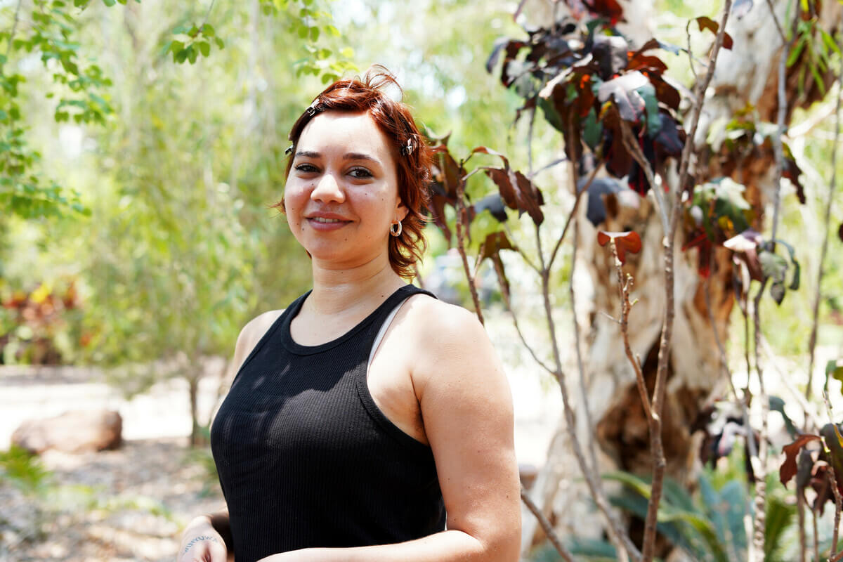 Obby stands smiling at the camera in front of some trees in Yawuru (Broome), Australia. Obby participated in the KAWC Roundtable in Broome, 2023. She is a proud Bunuba woman from Fitzroy Crossing.