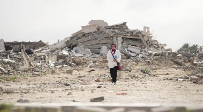 Gaza, Palestinian Territory, Occupied: Fidaa Shurrab, Director of Projects at Atfaluna for Deaf Society in Rafah stands in front of a destroyed building in Gaza strip while going to work. Photo: Alef Multimedia/Oxfam