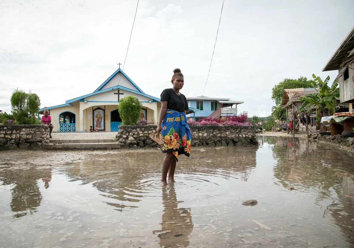 Lilisiana, Malaita Province, Solomon Islands: Grace Namoi (19), a student at the local high school, walks through water left in the town square after a high tide. Grace worries about the future of her town with the visible impacts of a changing climate. Lilisiana, Malaita Province, Solomon Islands: Grace Namoi (19), a student at the local high school, walks through water left in the town square after a high tide. Grace worries about the future of her town with the visible impacts of a changing climate.