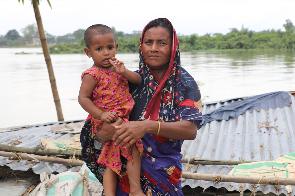 Bangladesh: Khadija Akhtar, 33 year old, (stands with her child) in front of her submerged house. She was displaced after flood in Sylhet and Sunamganj region in 2022. Photo: Mutasim Billah/Oxfam